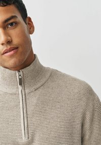 Jonge man in een beige geribbelde trui met een half-rits kraag, getoond vanaf de schouders omhoog tegen een effen witte achtergrond.