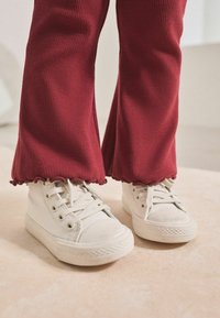 Baskets blanches en toile avec des lacets plats et des semelles en caoutchouc, associées à un pantalon rouge à côtes évasé avec un ourlet en feston.