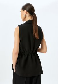 Blusa nera senza maniche con vita arricciata, tessuto morbido e colletto strutturato. Presenta un design pulito e tasche su entrambi i lati.