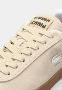 Baskets en daim beige avec lacets crème, semelle marron et logo crocodile blanc sur le côté, présentant des détails cousus et une étiquette de languette marquée.