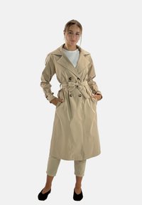 Beige trenchcoat med dubbelknäppt knäppning, ett knytbälte och stora slag, bärs över en vit topp och ljusgröna byxor.