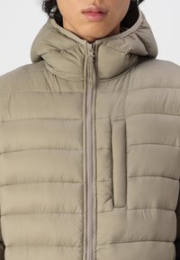 Béžová prošívaná bunda s kapucí, se středním zipem, vodorovným prošíváním a malou kapsou na hrudi. Měkká textura, lehký design.