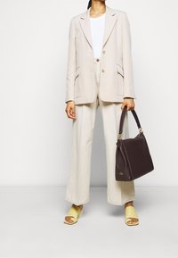 Person iført en beige blazer, hvid bluse, brede cremede bukser, gule åbne hæle og holder en mørkebrun lædertaske i højre hånd.