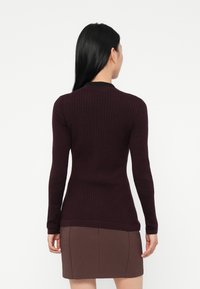 Woman with long black hair wearing a fitted dark purple ribbed sweater and a brown skirt, facing away against a white background.