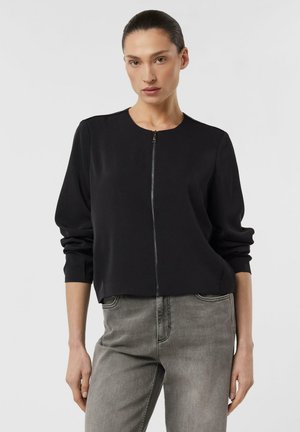 Kobieta w czarnej bluzie na zamek z długim rękawem i wyblakłych szarych dżinsach, stojąca na tle jasnego, jednolitego tła.