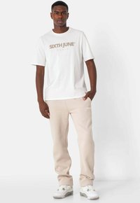Hvid bomulds T-shirt med "SIXTH JUNE" logo i beige. Beige sweatpants med sidelommer. Hvide sneakers med grå accenter. Glat tekstur.