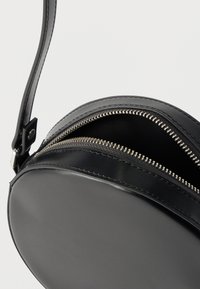 Στρογγυλή μαύρη δερμάτινη τσάντα με λεπτό λουρί, με ασημένια φερμουάρ και λεία υφή. Δύο εμφανή χωρίσματα.