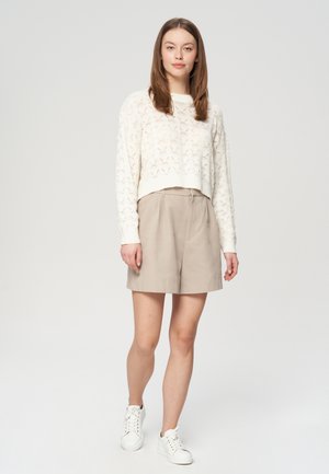 Kremowy, dziany sweter w krótkim kroju z otwartym wzorem, zestawiony z beżowymi, dopasowanymi szortami i białymi trampkami. Prosta, nowoczesna sylwetka o luźnym kroju.