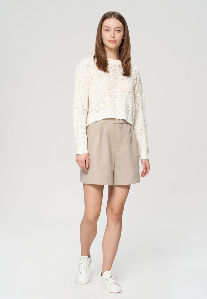 Kremowy, dziany sweter w krótkim kroju z otwartym wzorem, zestawiony z beżowymi, dopasowanymi szortami i białymi trampkami. Prosta, nowoczesna sylwetka o luźnym kroju.