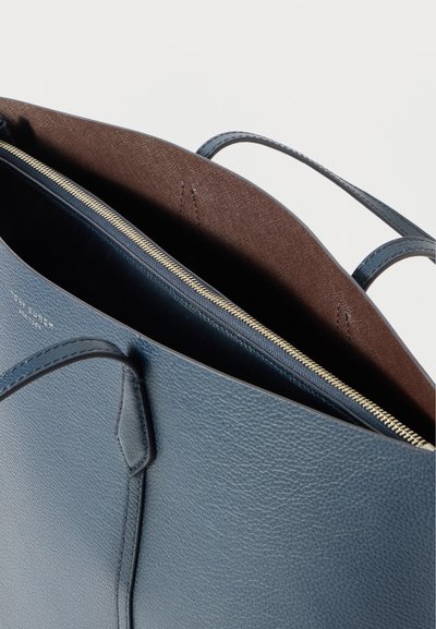 Bolsa tote de couro azul com textura suave, apresentando um interior de tecido castanho e um fecho de zíper dourado. Alças curtas para fácil transporte.