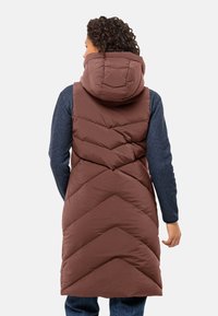 Bruine puffer vest met een capuchon, met een chevron quiltpatroon. Gemaakt van een zachte stof, gedragen over een blauwe langwerpige top.