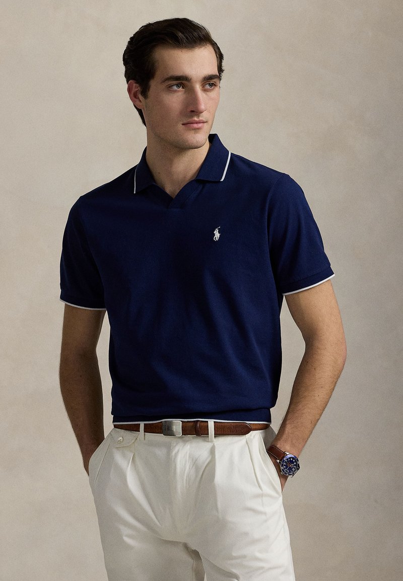 Πόλο μπλε ναυτικού με λευκή επένδυση στον γιακά και τα μανίκια, με μικρό λογότυπο στο στήθος, συνοδευόμενο από ανοιχτόχρωμο παντελόνι και δερμάτινη ζώνη.