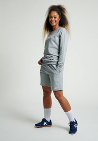 Felpa grigia e pantaloni corti realizzati in un materiale morbido, caratterizzati da una vestibilità rilassata. Scarpe sportive blu e calzini bianchi con dettagli neri completano l'outfit.
