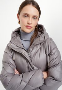 Šedá prošívaná bunda s kapucí, s podrobným prošíváním a lesklým povrchem. Nositá přes světle šedý rolák, který má jemné kovové akcenty.