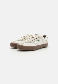 Scarpe da ginnastica bianche in tela con suole in gomma marrone, punta arrotondata, design con lacci e strisce laterali. Accento verde del logo vicino al tallone.