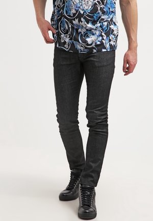 Hombre con jeans negros ajustados, zapatillas negras de caña alta y una camisa negra con un patrón floral azul y blanco, de pie frente a un fondo blanco.