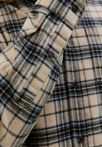 Beige Flanellhemd mit schwarzem und blauem Karomuster. Enthält Knopfdetails an den Ärmelbündchen und hat eine weiche, strukturierte Stoffoberfläche.