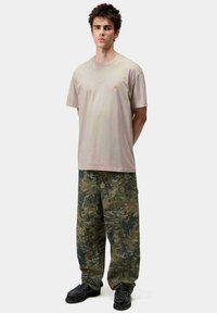 Camiseta de algodón beige con un pequeño logo en el pecho, combinada con pantalones cargo holgados de camuflaje en tonos verdes y marrones. Zapatillas negras.