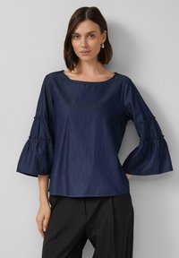 Navyblaues Denim-Oberteil mit weit ausgeschnittenem Hals, drei Viertel langen Glockenärmeln mit Raffung und lockerer Passform. Kombiniert mit schwarzen Hosen.