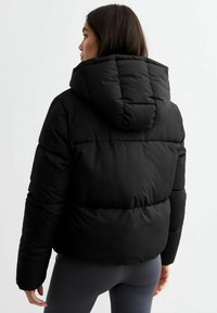 Kobieta w czarnej kurtce puchowej z kapturem, odwrócona plecami, w ciemnoszarych legginsach na jednolitym białym tle.