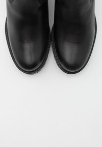 Botines de cuero negro con acabado liso, punta redonda y detalles de costura alrededor de la suela. Colocados uno al lado del otro sobre un fondo blanco.