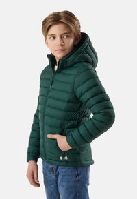 Garçon portant une veste matelassée à capuche verte foncée avec des poches zippées, regardant sur le côté, debout devant un fond blanc.