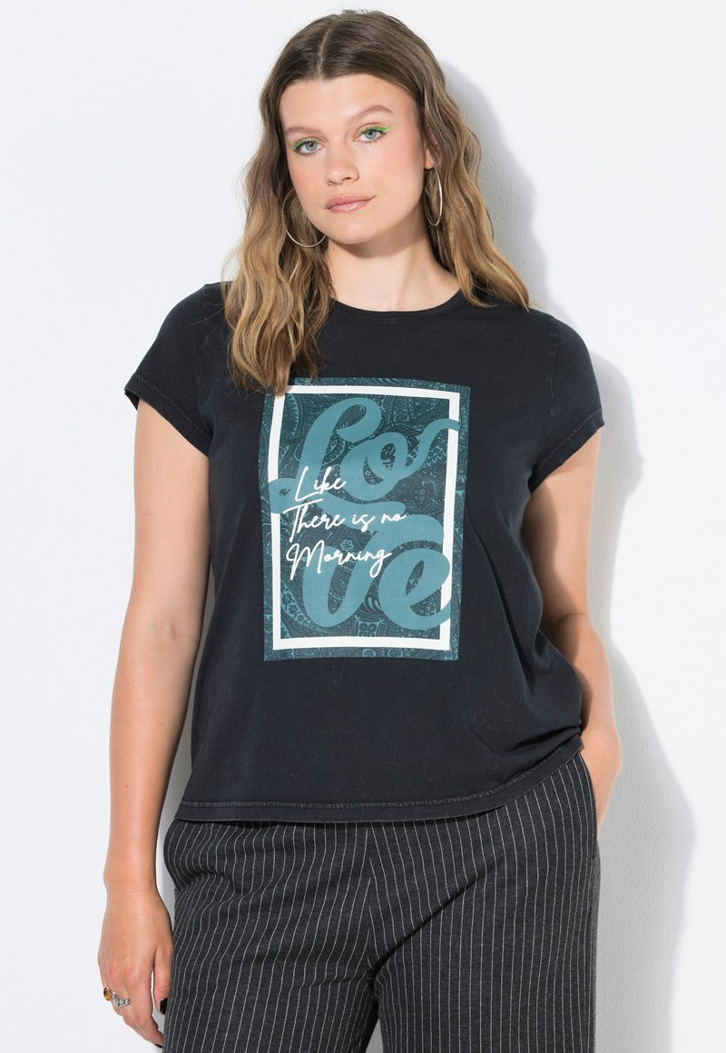 Mujer con cabello largo y ondulado, que lleva pendientes de aro negros, una camiseta negra con el texto "Ama como si no hubiera mañana" y pantalones a rayas.