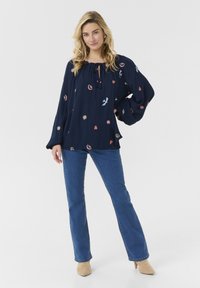 Blouse bleu marine avec des motifs brodés en rose, jaune et blanc. Il a un col à cordon de serrage et des manches larges. Associée à un jean bleu.