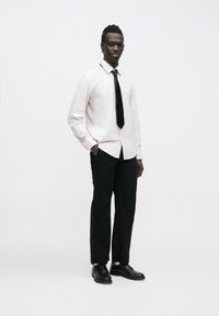 Mladý muž stojaci s rukami v klopách, oblečený v ľahkej pruhovanej košeli, čiernej kravate, čiernych nohaviciach a čiernych spoločenských topánkach na bielom pozadí.