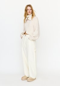 Kobieta z rozpuszczonym blond włosami, ubrana w kremową, miękką kurtkę na zamek, białe spodnie o szerokich nogawkach z boczną kieszenią oraz beżowe buty na platformie.