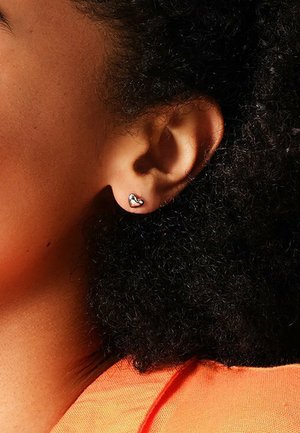 Orecchino a perno a forma di cuore in argento su un orecchio scuro, che mette in evidenza la superficie metallica liscia contro i capelli ricci e texturizzati.