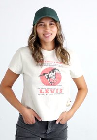 Kremfarget bomulls t-skjorte med rød grafikk av en løpende hest og "LEVI'S"-tekst. Kombinert med en mørkegrønn baseballcaps og grå jeans.