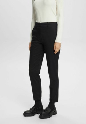 Kobieta ubrana w czarne, wysokie spodnie cygaretki, czarne mokasyny na grubych podeszwach, czarne skarpetki oraz biały, prążkowany, długi rękaw bluzki.