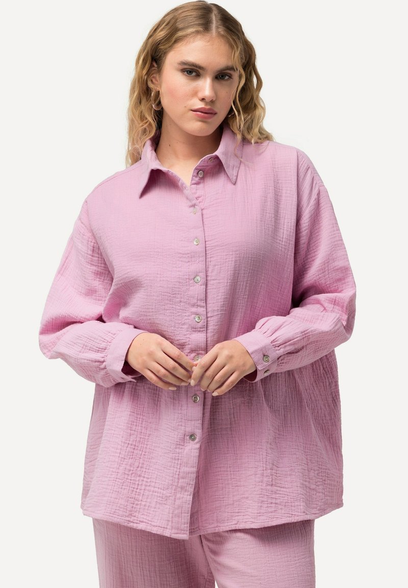 Jasnoróżowa, oversize koszula z guzikami, wykonana z teksturowanej tkaniny, z kołnierzykiem i dużymi rękawami. Modelka pozuje z dłońmi złożonymi razem.