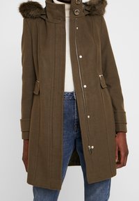 Mujer vistiendo un abrigo de lana verde oliva con capucha con borde de piel, cierre de cremallera y botones a presión, combinado con jeans azules y un suéter blanco de cuello alto.