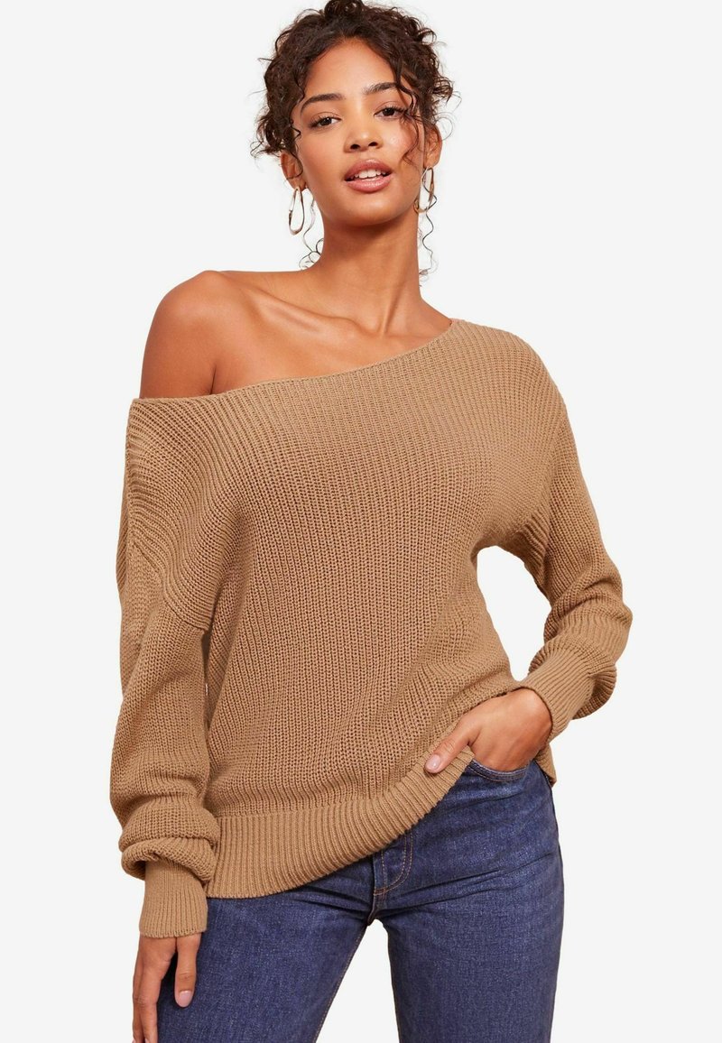 Strickpullover in Beige mit einem schulterfreien Design, weiten Ärmeln, geripptem Textur und einer lässigen Passform, kombiniert mit dunklen Jeans.