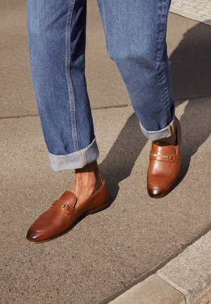 Mocassins en cuir marron au design épuré, à bout rond et accents de boucles en laiton, assortis à un jean en denim bleu retroussé.