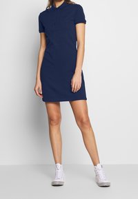Abito polo blu navy con maniche corte, forma aderente, colletto con tre bottoni e logo ricamato sul petto. Indossato con sneakers alte bianche.