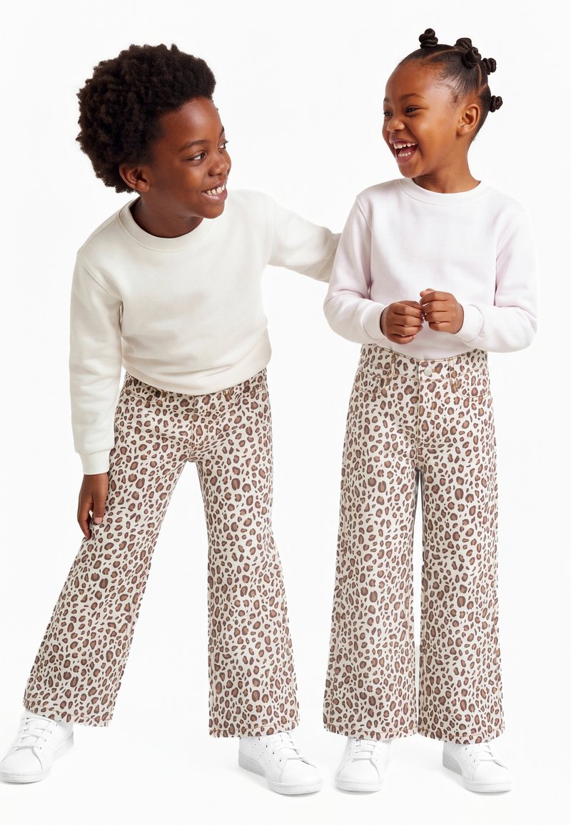 Deux enfants souriants portant des hauts blancs assortis, des pantalons larges à imprimé léopard et des baskets blanches sur fond blanc.