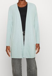 Hellblauer Cardigan aus weichem Material, mit offenem Design und langen Ärmeln, ausgestattet mit zwei Seitentaschen, über einem schwarzen Oberteil getragen.