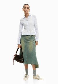 White, textured button-up shirt with peplum waist. Long, green denim skirt, ankle-length. Model holds a brown bag, wearing white sneakers.