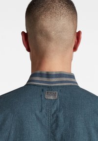 Chaqueta de mezclilla azul oscuro con un cuello rayado. Presenta una etiqueta en la parte posterior y un diseño simple y limpio. La tela tiene un acabado texturizado.