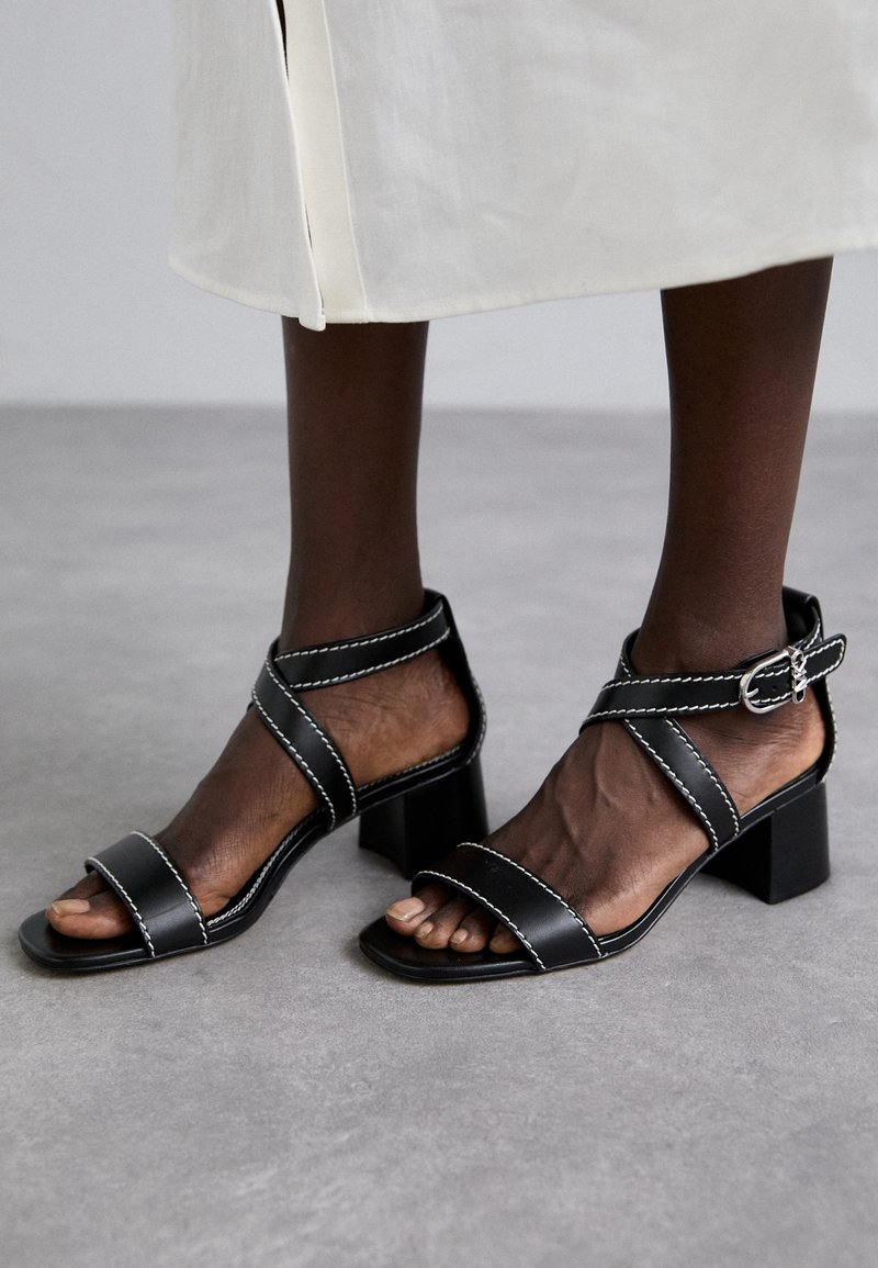 Czarne sandały na blokowym obcasie mają dwa szerokie paski, krzyżującą się opaskę na kostce oraz białe detale przeszycia. Wykonane są z gładkiej skóry.