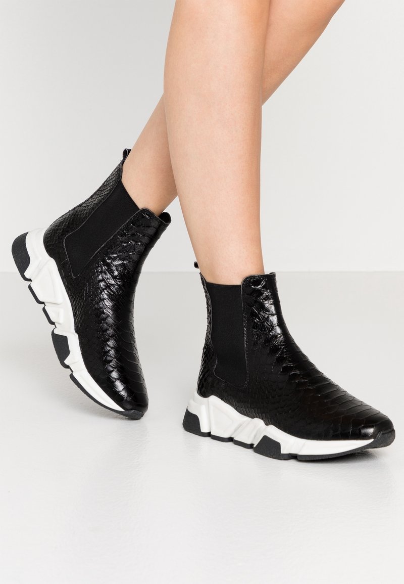 Bottes noires à la cheville avec motif texturé, semelles épaisses blanches, panneaux latéraux élastiques, portées sur des jambes croisées sur un fond clair uni.