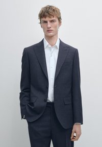 Abito blu navy scuro in tessuto liscio, design monopetto con due bottoni, abbinato a una camicia bianca impeccabile e pantaloni slim fit.