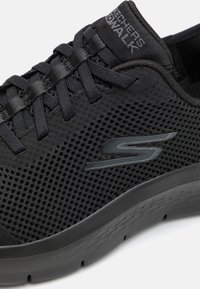 Sneaker noire avec une tige en maille, des accents texturés, des lacets plats et une semelle texturée. Présente un logo en finition brillante sur le côté.
