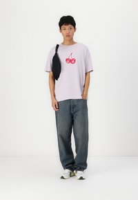Paarse katoenen t-shirt met een rode kers graphic, gecombineerd met blauwe spijkerbroek en witte sneakers met groene accenten. Zwart schoudertasje.