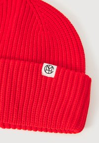 Red knit beanie with a ribbed texture and a folded cuff. Features a small white tag with a black logo on the side.