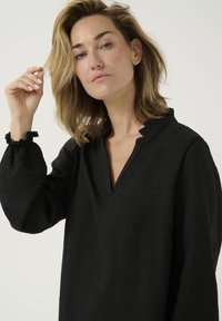 Black, long-sleeve blouse with a scalloped neckline and gathered details at the cuffs. Made from a textured fabric.