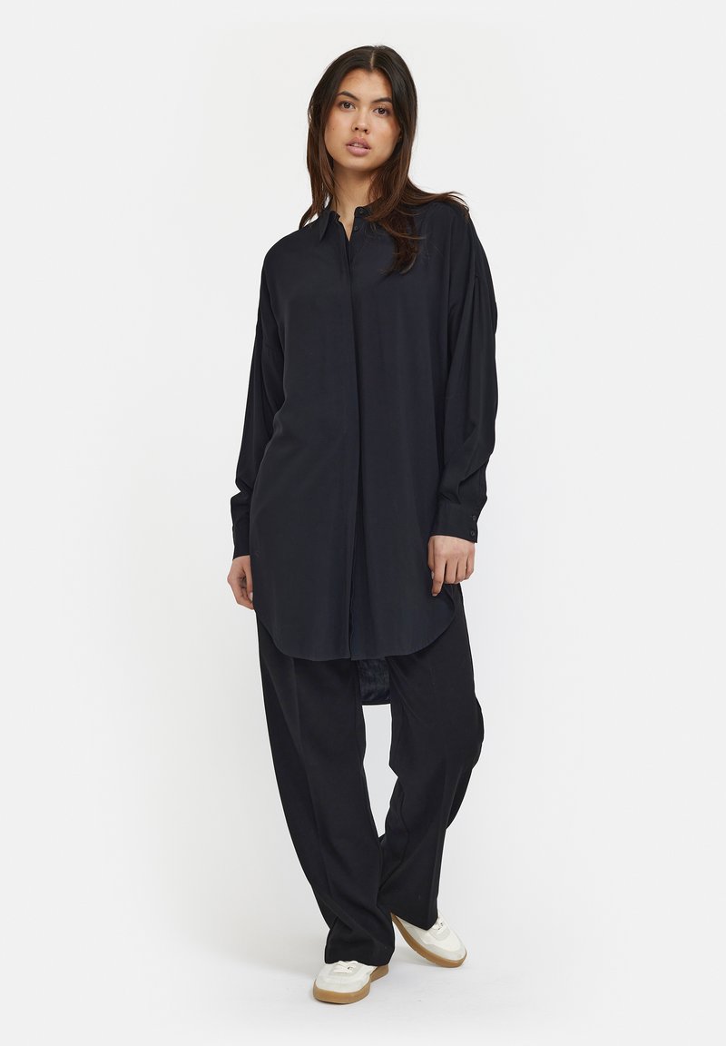 Chemise noire à manches longues avec un col boutonné, texture lisse et ourlet arrondi, associée à un pantalon noir ample et des baskets blanches.